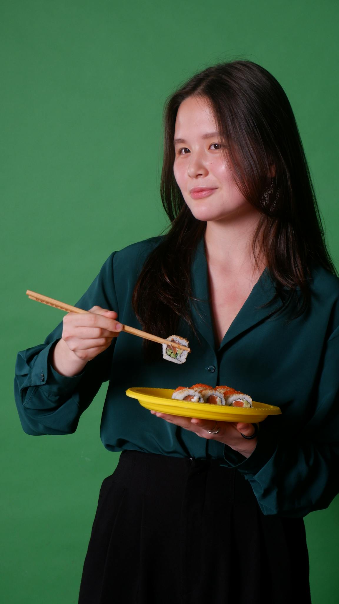 Portrait of an Asian woman holding sushi with chopsticks against a green background.